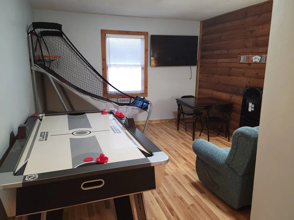 A vibrant game room featuring an air hockey table, basketball arcade game, flat-screen TV, dining table, and blue chair. The setup exudes a lively, family-friendly atmosphere perfect for friendly competitions and movie nights.