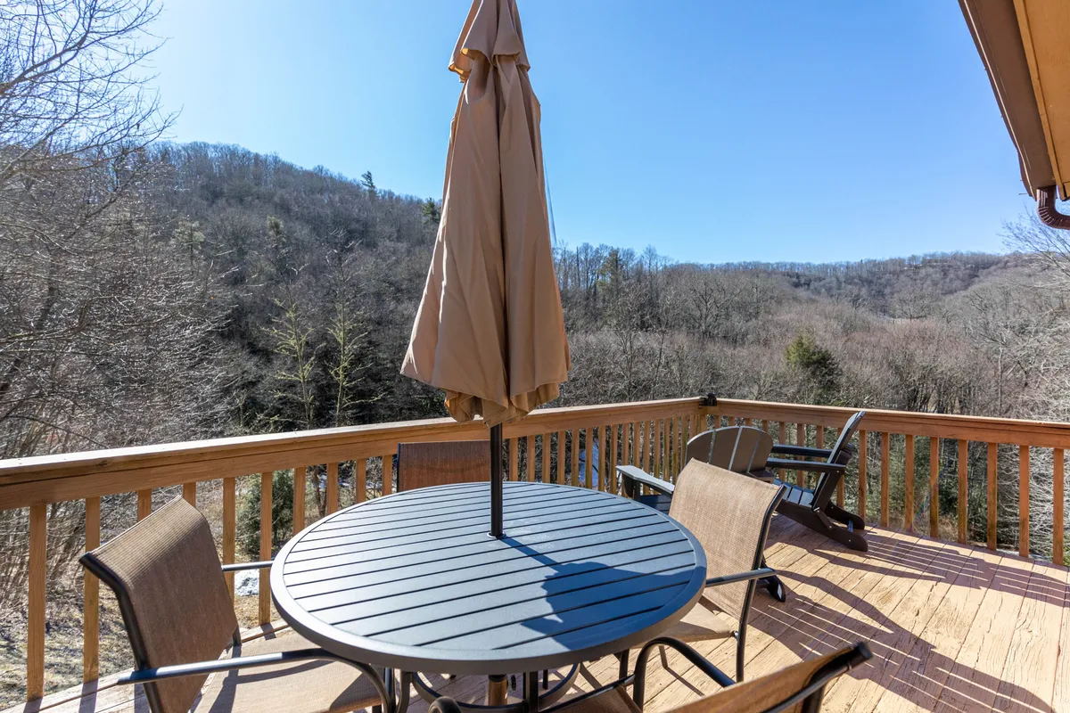 A wooden deck featuring a table with an umbrella, surrounded by lush trees and offering sweeping mountainous views. The scene suggests a peaceful spot for outdoor enjoyment, with nature as the backdrop and ample room for socializing or quiet reflection.