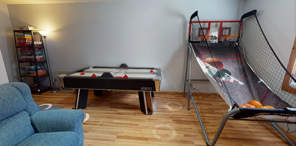 Another angle of the game room highlighting the air hockey table, basketball shooting game, a comfortable chair, and bookshelf. The room feels inviting, playful, and full of entertainment options.