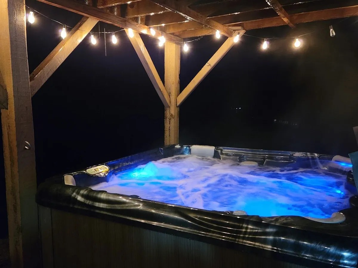 The image showcases the exterior of Pine Creek Cabin at dusk, with a warm wooden façade softly illuminated by charming string lights. The inviting hot tub sits in the foreground, steaming gently and promising serene relaxation. Lush greenery surrounds the cabin, and the scene evokes an atmosphere of tranquility and togetherness, perfect for unwinding or stargazing with friends and family.