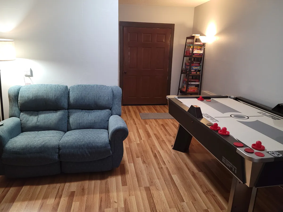 Interior view of the game room with a sofa, air hockey table, and bookshelf. The scene suggests relaxed play, socializing, and multiple entertainment options. The air hockey table is a focal point, aligning perfectly with the activities described in the section.