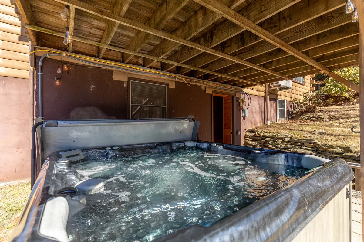 A private outdoor hot tub nestled beside the cabin, framed by sturdy wooden beams and adjacent to the exterior wall. The scene captures the inviting warmth of the bubbling water, with hints of surrounding greenery and a sense of seclusion. The space is clearly designed for relaxation and gathering, offering views of the outdoors.