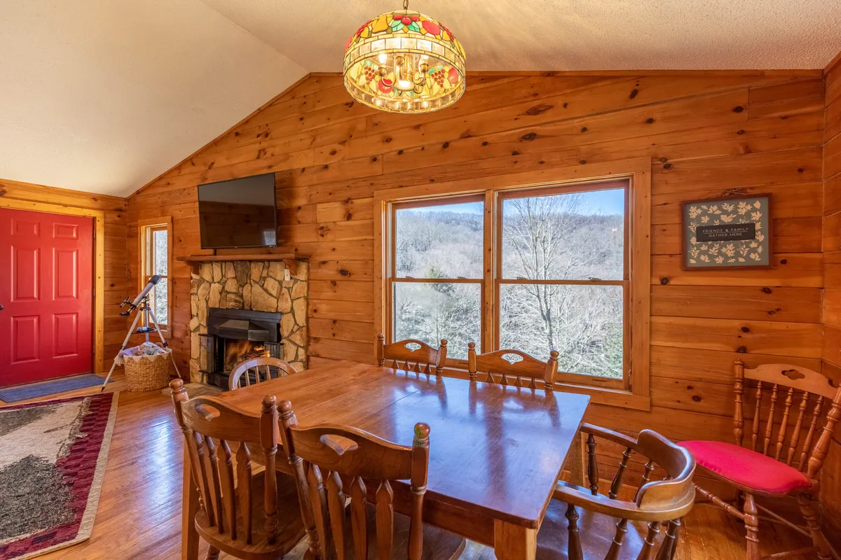 A cozy dining area with a large wooden table set for six, a colorful chandelier overhead, and a fireplace in the background. Large windows offer scenic views, making the space perfect for lingering meals, game nights, and fireside chats.