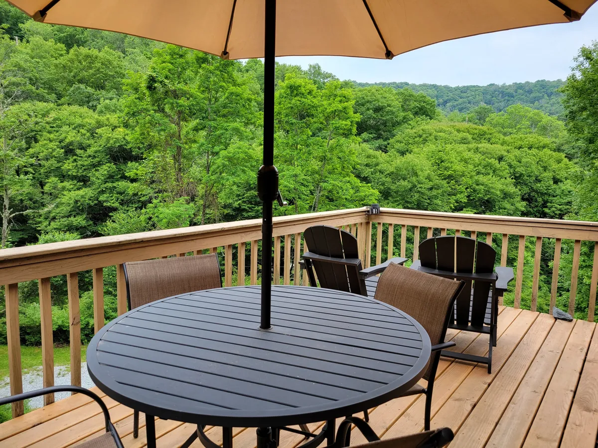 An inviting wooden deck surrounded by lush greenery, featuring a table with an umbrella that offers a shady spot to relax or dine outdoors. The setting is private and immersed in nature, with no neighboring homes in sight, creating a tranquil and secluded atmosphere ideal for outdoor activities and enjoying fresh mountain air.