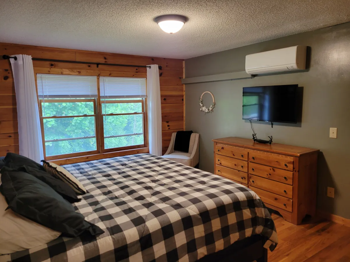 This image depicts a comfortable bedroom with plaid bedding, a large window letting in natural light, a wooden dresser, and a TV mounted on the wall. The warm wood tones and inviting decor create a serene atmosphere, perfectly suited for restful sleep.
