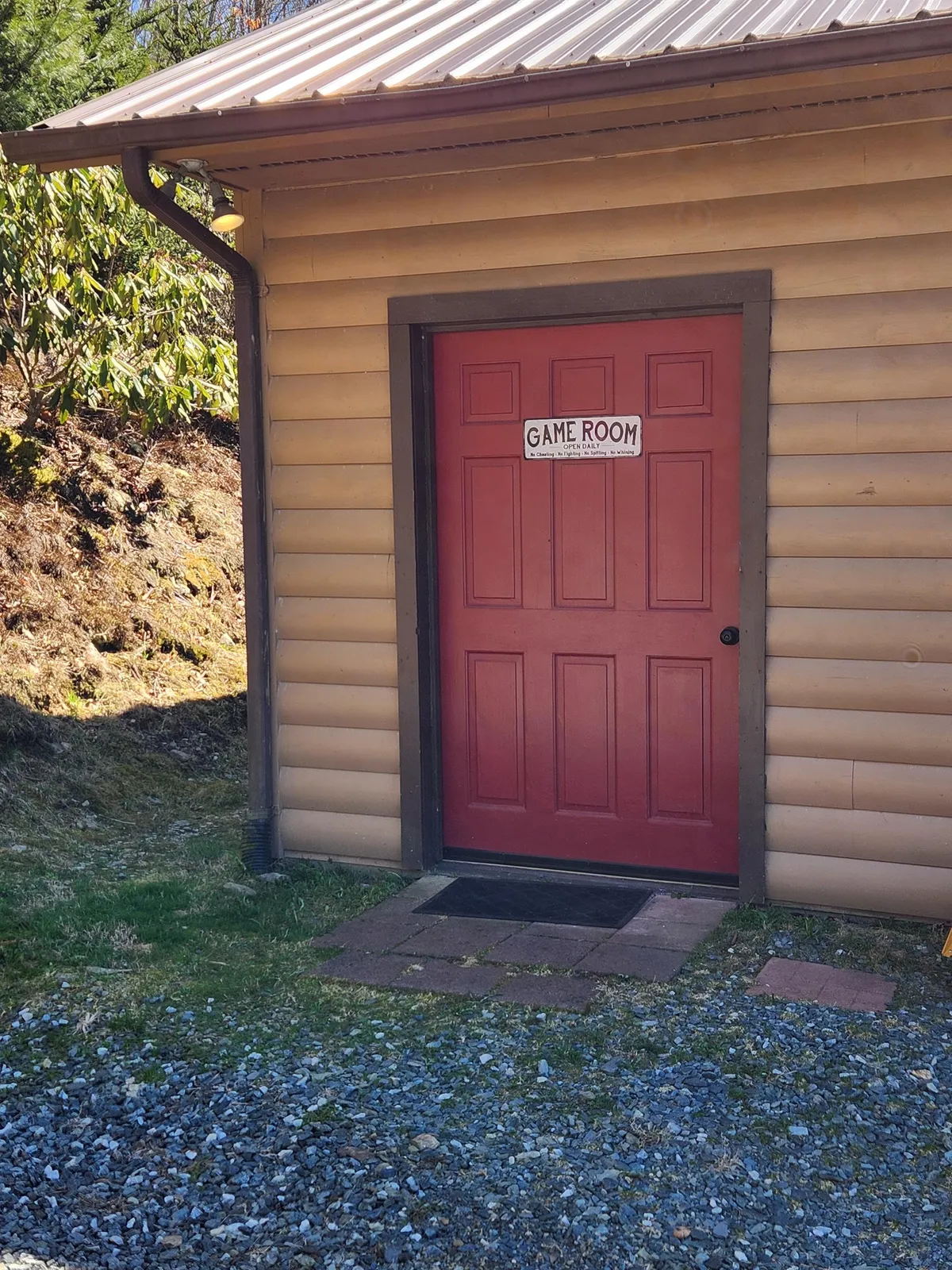 Another exterior view of the game room with a red door, a clear game room sign, wooden siding, and lush greenery. The image conveys a playful and inviting mood, highlighting the unique, dedicated recreation space within the property’s natural setting.