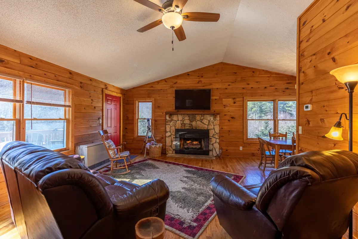 A warmly lit living room featuring a striking stone fireplace as the centerpiece, surrounded by plush, comfortable seating. The wood paneling and large windows fill the room with natural light and offer peaceful wooded views, creating an atmosphere that feels both cozy and connected to the outdoors. A smart TV is visible, blending modern convenience with rustic charm.