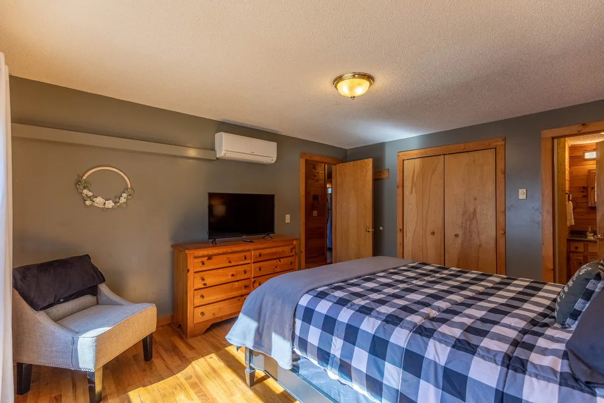 A cozy bedroom featuring a bed with checkered bedding, a wooden dresser, a TV, and an armchair tucked beside a window. The scene radiates warmth and comfort, ideal after a wintry outdoor adventure.