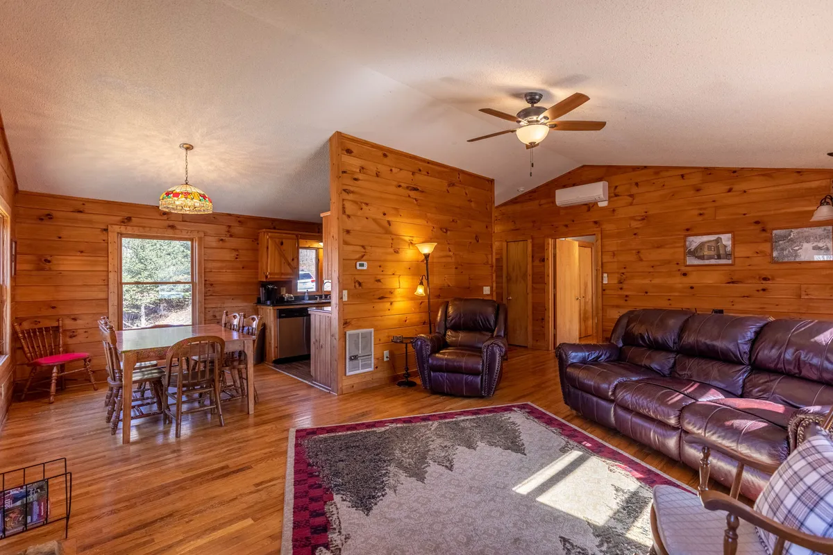 A bright, open-plan living and dining area with wooden walls, a comfortable leather sofa, a rustic dining table, and a rug featuring a mountain design. The space feels welcoming and perfect for family gatherings or relaxed conversations, with atmospheric lighting and a ceiling fan adding comfort and style.